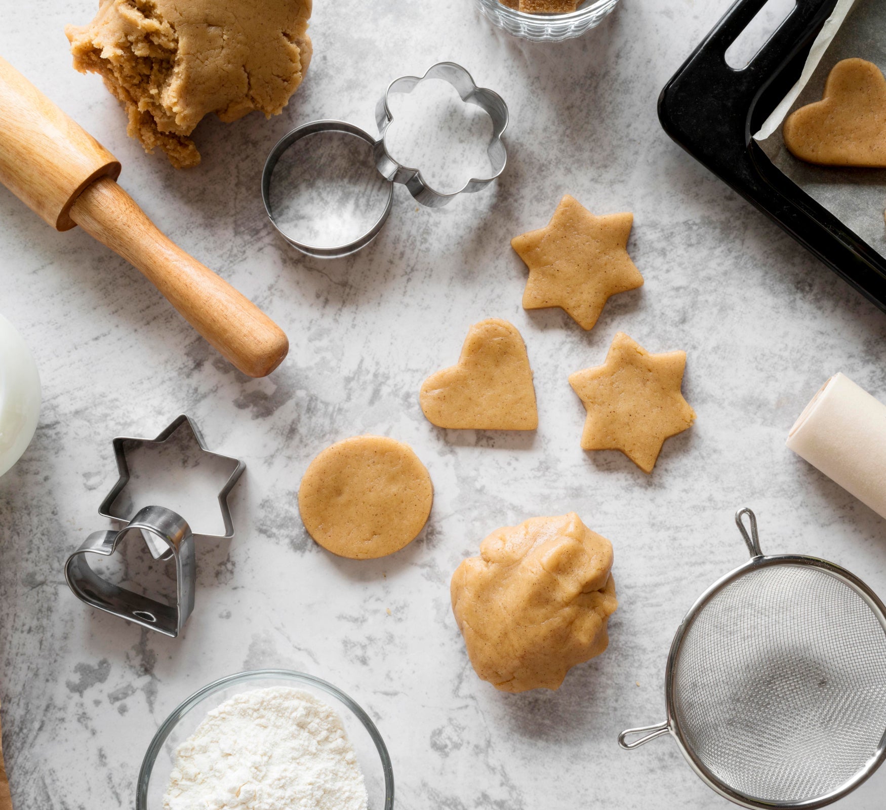 Assorted cookie cutters in various shapes, a wooden rolling pin, and freshly cut cookies displayed on a white table, highlighting essential tools for creative baking.
