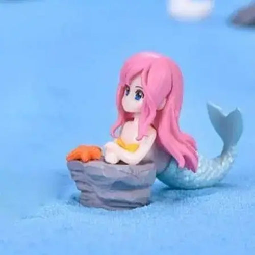 Miniature mermaid figurine with pink hair, yellow bikini top, and blue tail resting on a rock with an orange starfish – perfect for under the sea cake toppers, ocean theme party décor, and kids' birthday mermaid decorations.