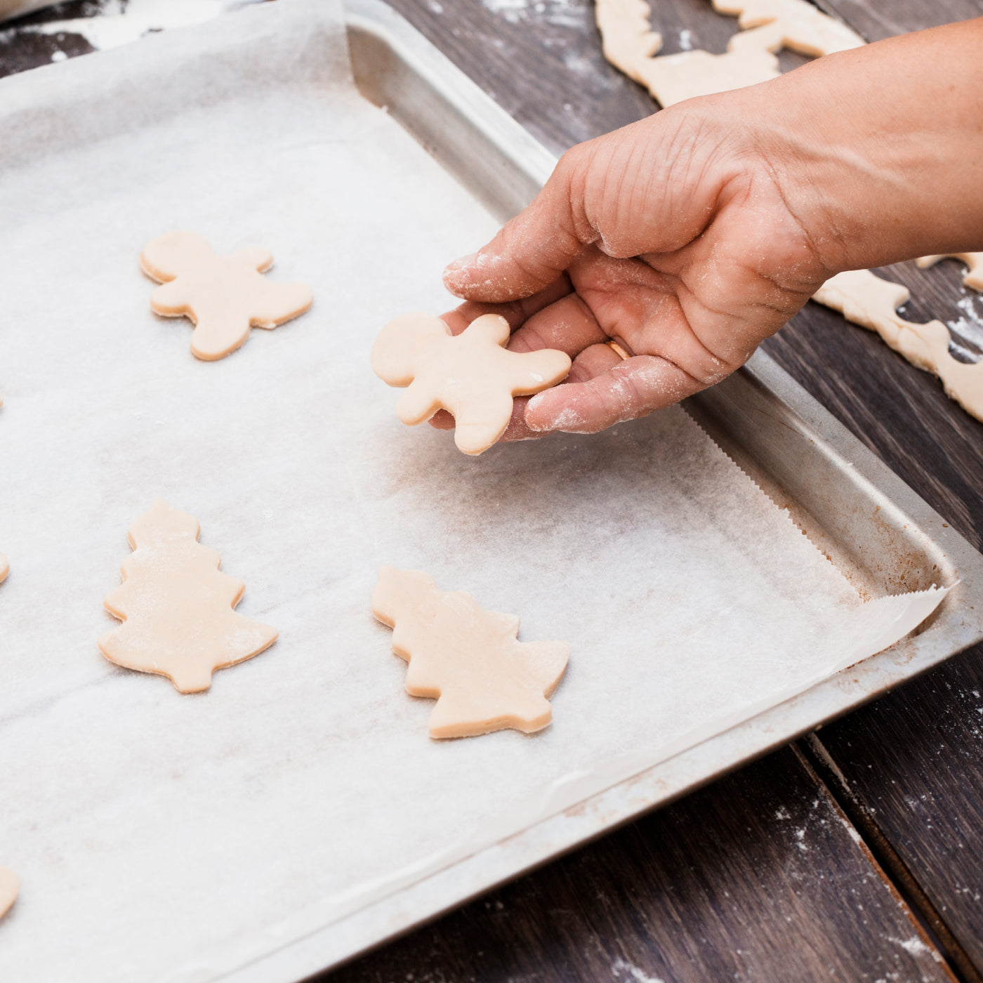 Hand placing raw Christmas-themed cookies including gingerbread men and trees on a baking sheet lined with parchment paper — festive holiday baking prep.