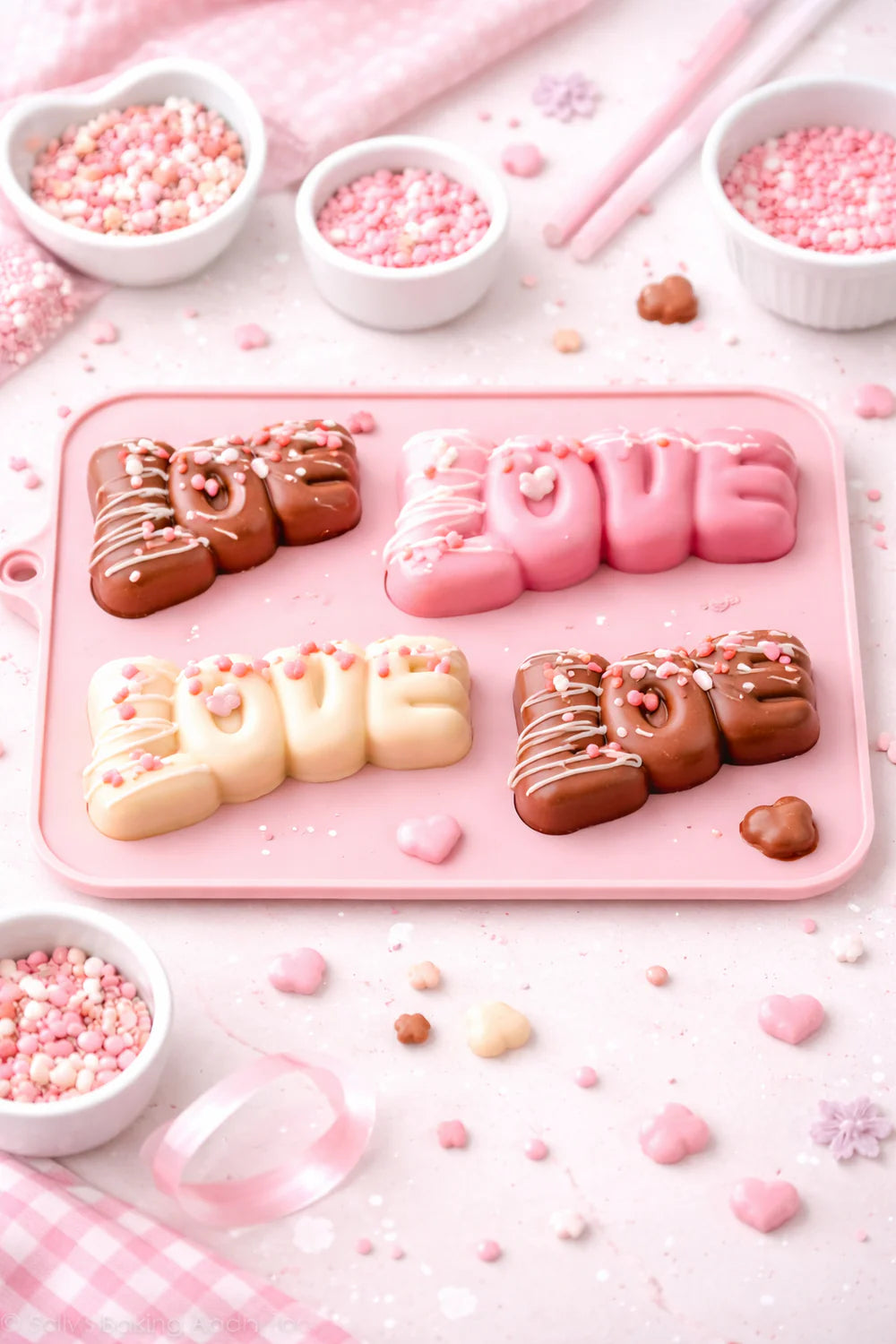Chocolate letters spelling 'LOVE' on a pink silicone chocolate mold with pink and white sprinkles around.
