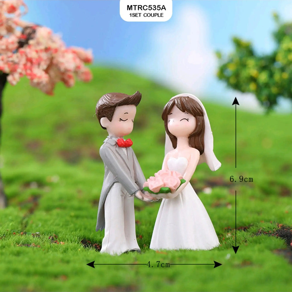 Wedding couple figurine with 6.9 cm height and 4.7 cm width – groom in grey suit with red bowtie holding pink flower bouquet with smiling bride in strapless white gown and veil outdoors