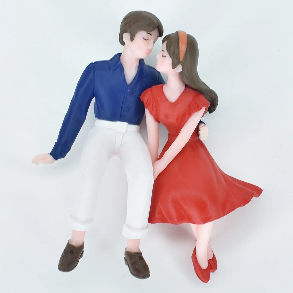 Miniature romantic couple figurine – boy in blue shirt and white pants sitting beside girl in red dress, perfect for love-themed cake toppers, anniversary gifts, and Valentine's decoration ideas.