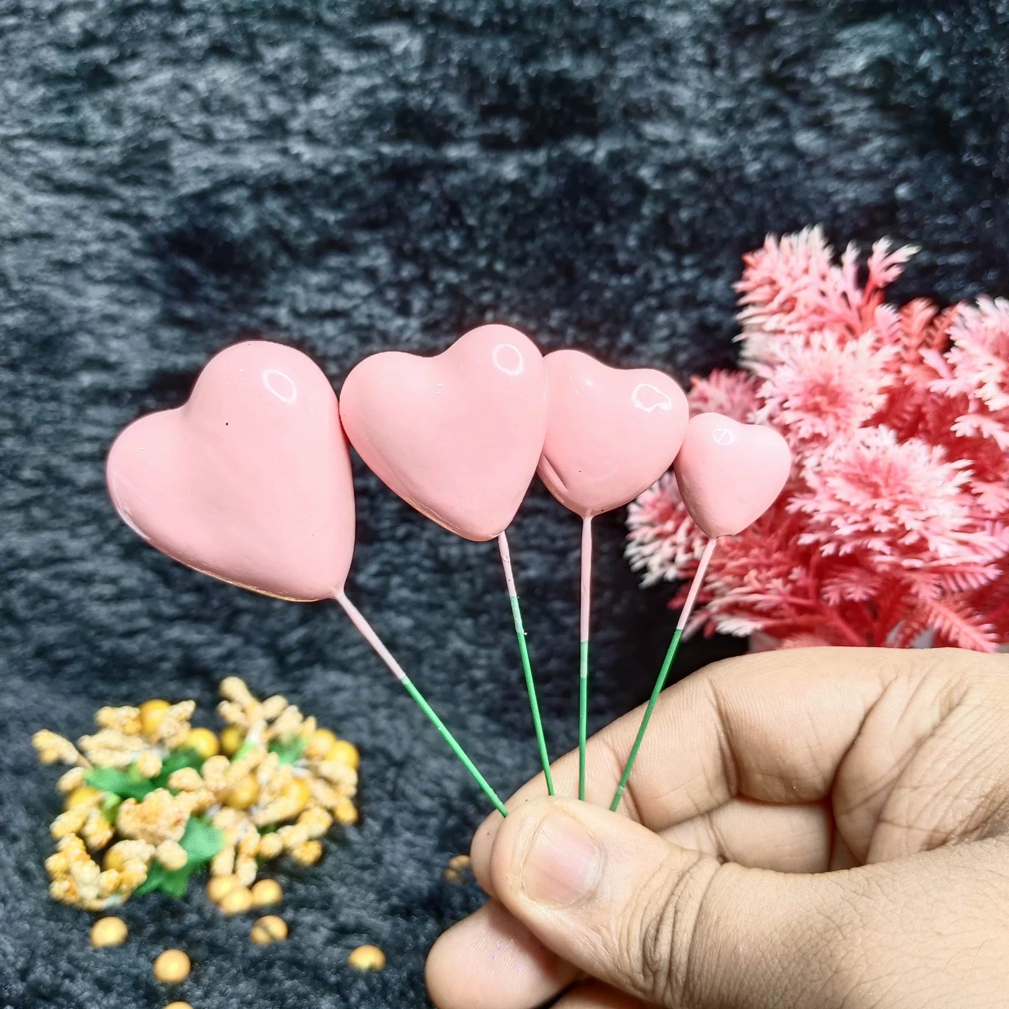 Heart-shaped pink faux balls held by a hand with a dark background and small plant.