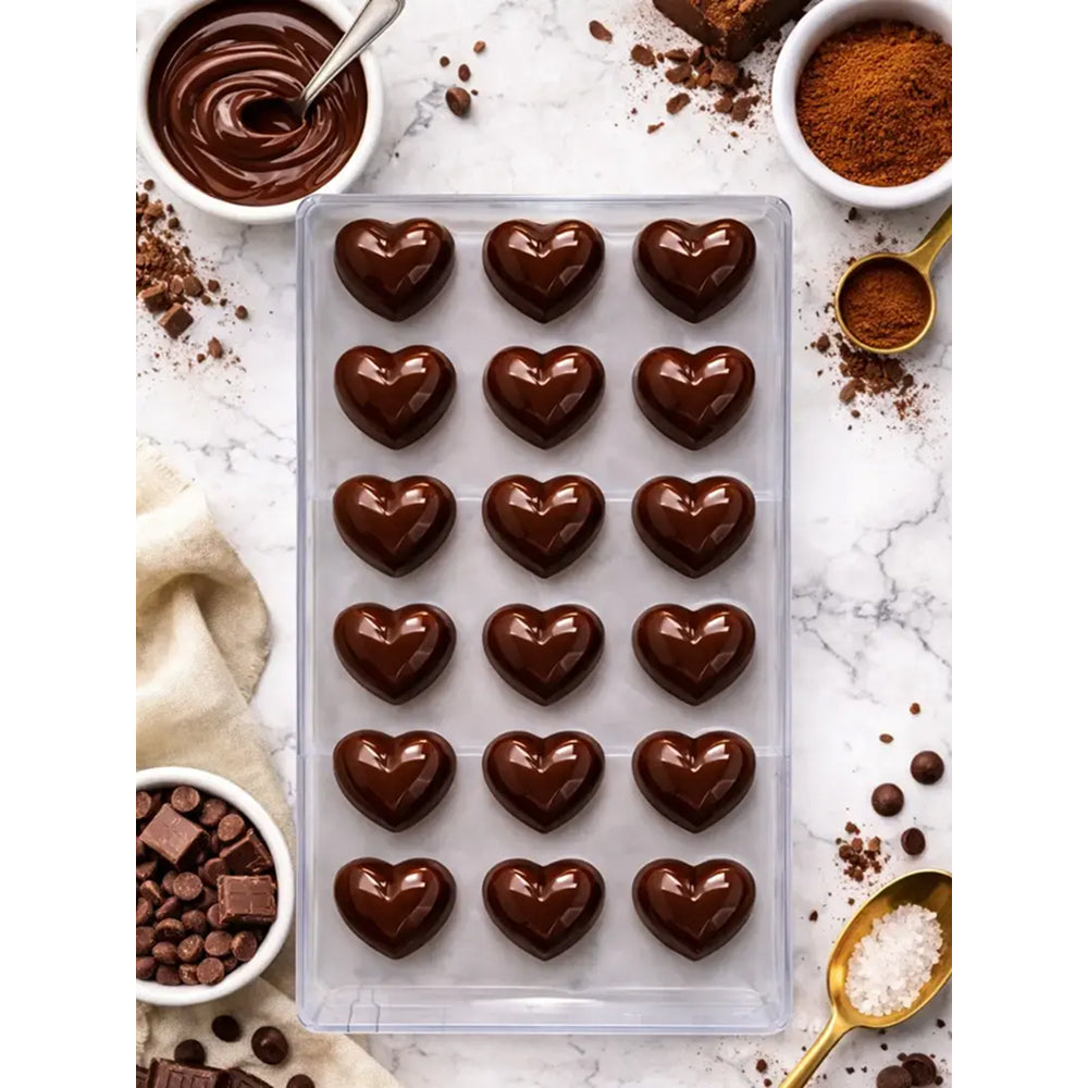 Heart-shaped polycarbonate chocolate candies in a mold surrounded by chocolate chips, cocoa powder, and a bowl of melted chocolate.