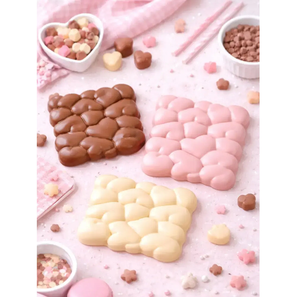 Three heart-shaped chocolate molds in brown, pink, and white on a pink background with small candies around.