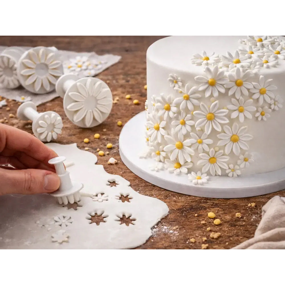 Decorative cake with white flowers and yellow centers on a wooden table, surrounded by floral-shaped cookie cutters.