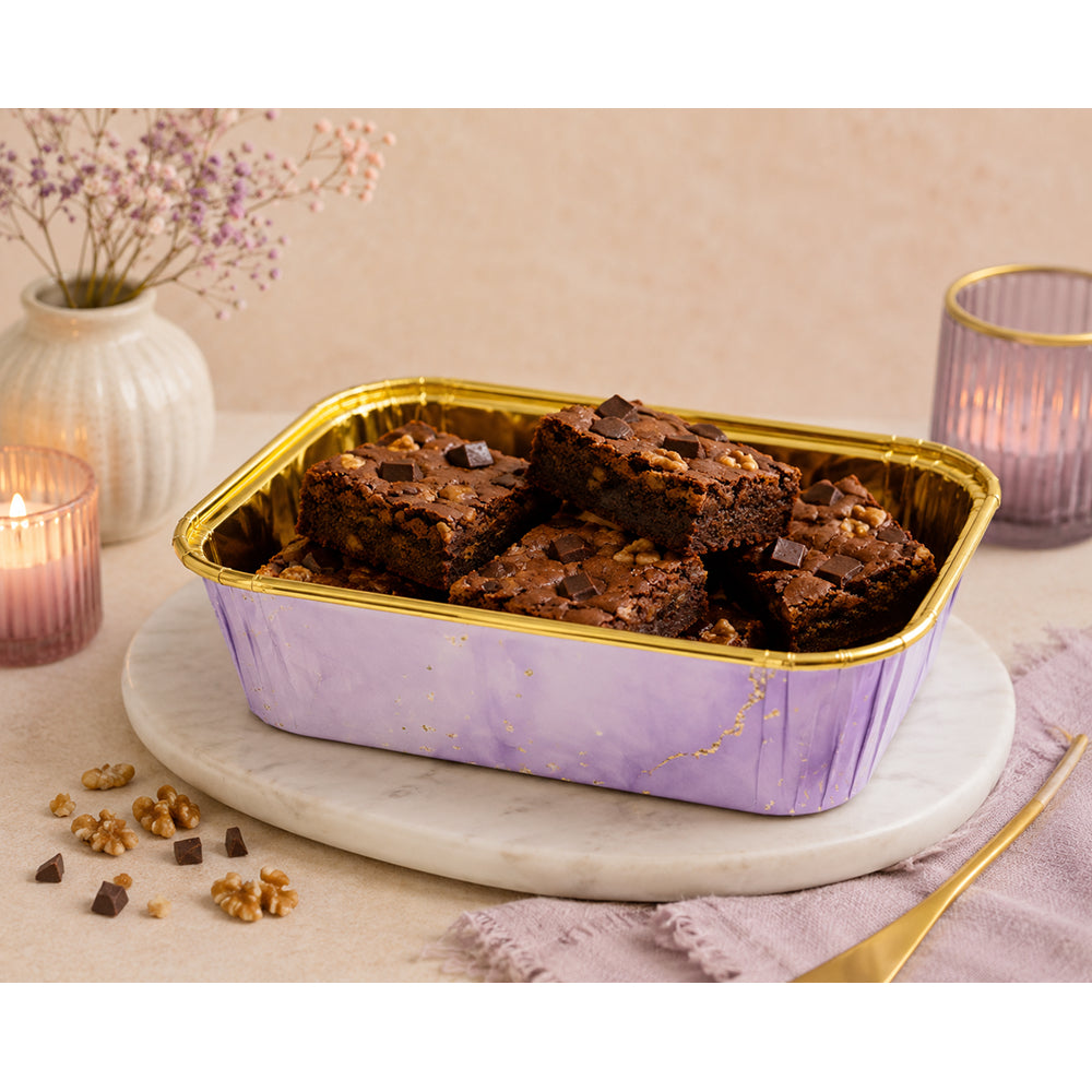 Square Muffin cup of brownies in a decorative purple and gold container on a marble surface with candles and flowers in the background.