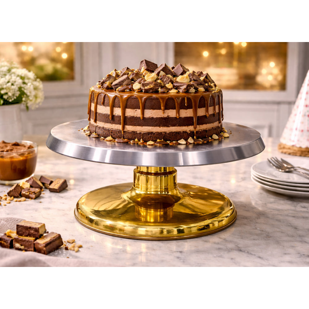 Chocolate cake on a gold cake stand with caramel sauce and chocolate pieces in a kitchen setting.