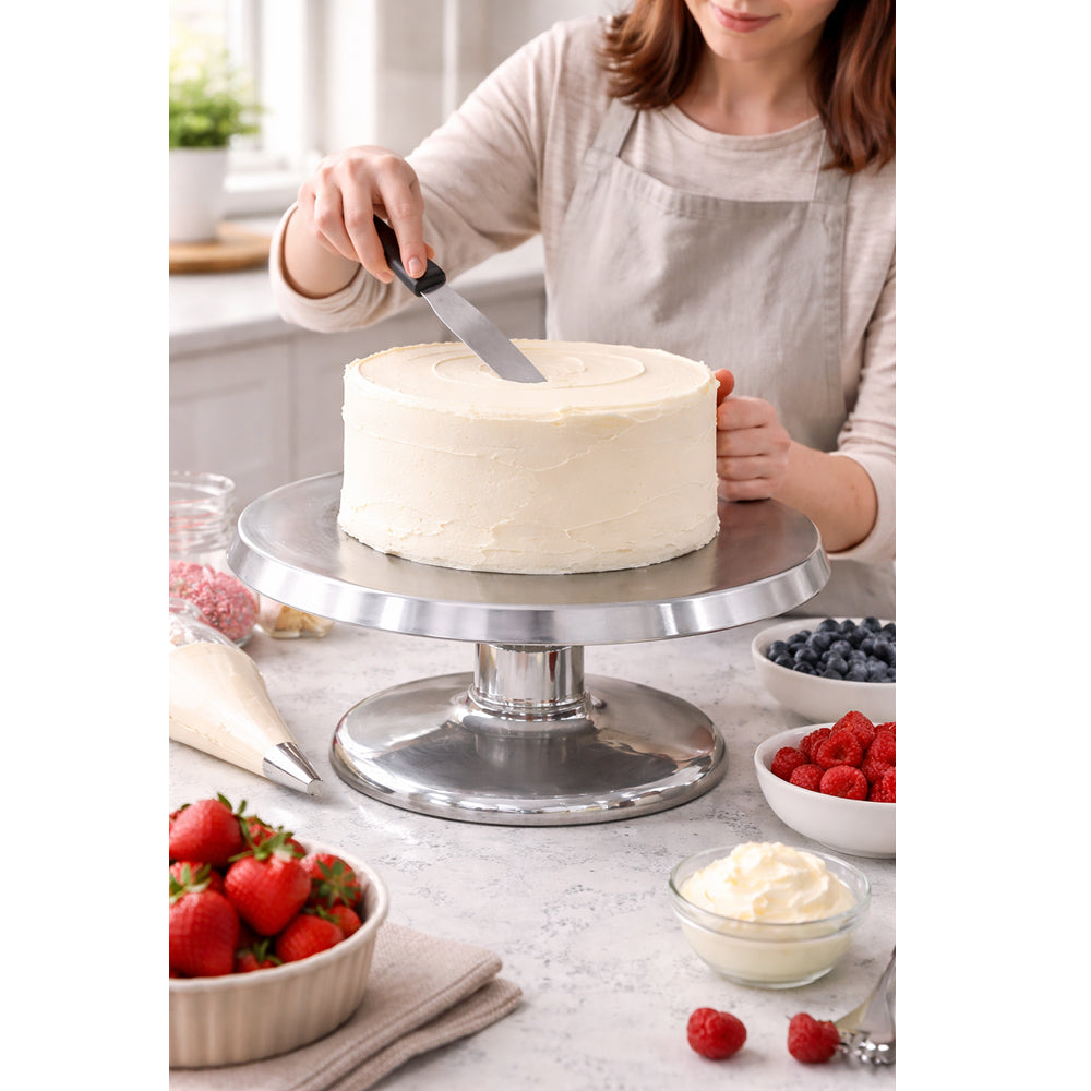 Person frosting a cake on a metal cake stand with various fruits and ingredients around.