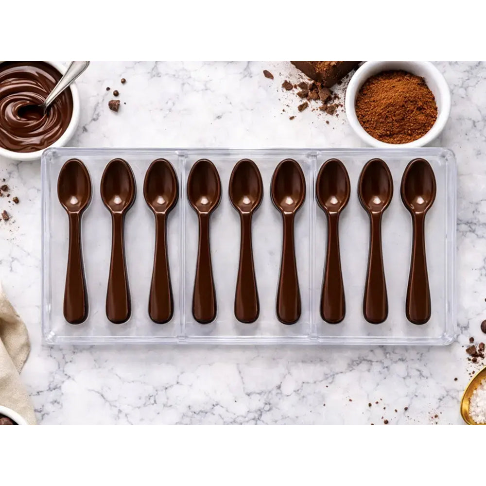 Chocolate-dipped spoons in a Polycarbonate tray on a marble surface with chocolate and cocoa powder.