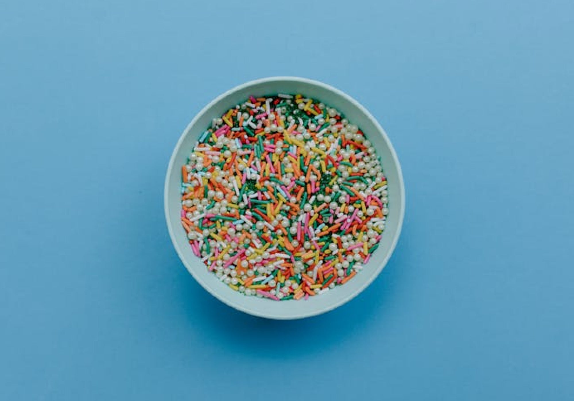 Top view of white bowl filled with vibrant multi-colored rainbow sprinkles placed on a solid blue background – minimal style image for baking decor themes and sweet treats.